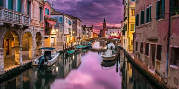 Un giorno a Venezia: canale di Venezia con colore viola del cielo che si riflette sull'acqua del canale. Le caratteristiche case affacciate sulla laguna sono illuminate dai lampioni e dalle luci del tramonto.
